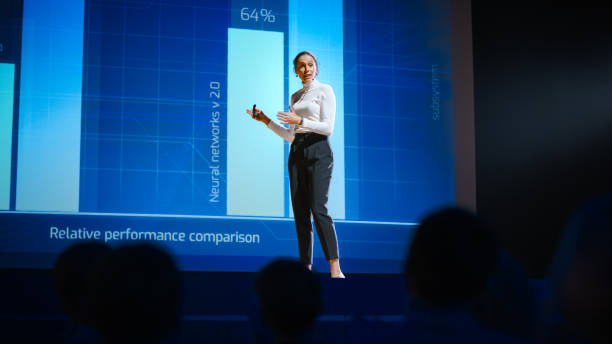 istock On Stage, Successful Female Speaker Presents Technological Product, Uses Remote Control for Presentation, Showing Infographics, Statistics Animation on Screen. Live Event / Device Release. 1226991539
