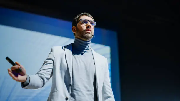 Photo of On-Stage Successful Male Speaker Presents Technological Product, Uses Remote Control for Presentation, Showing Infographics, Statistics Animation on Big Screen. Live Event / Business Conference