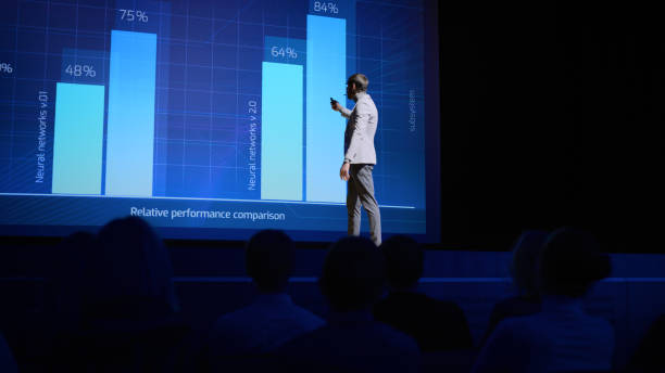 istock On-Stage: Speaker Does Presentation of the New Electronic Product, Shows Infographics, Statistic Animation on Screen. Auditorium Hall Live Event, Start-up Conference, Device Presentation and Release 1226991678