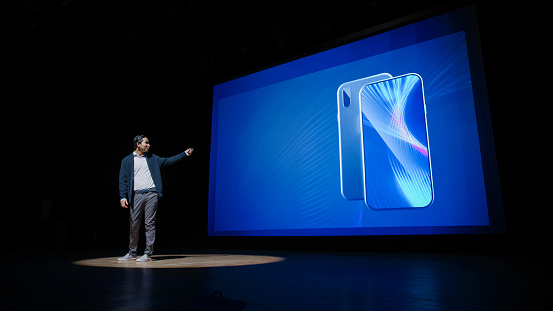 Live Event with Brand New Products Reveal: Speaker Presents Smartphone Device to Audience. Movie Theater Screen Shows Mock-up Touch Screen Mobile Phone with High-Tech Features and Top Highlights