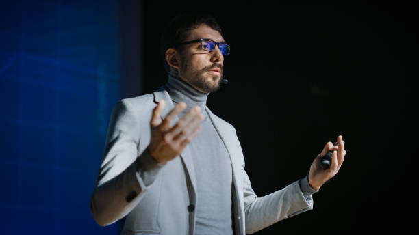 istock Great Motivational Speaker Stands on Stage, Talks about Happiness, Self, Success, Technology and How To Be Productive. Startup Presenter Pitching Ideas. Cinematographic Photo. 1226991765