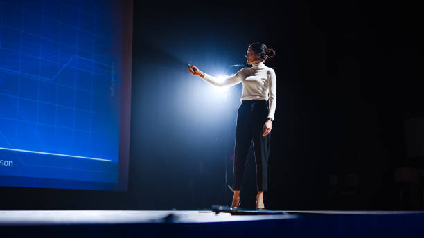 istock On-Stage Successful Female Speaker Presents Technological Product, Uses Remote Control for Presentation, Showing Infographics, Statistics Animation on Screen. Live Event / Device Release. 1226991771