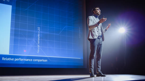 istock On-Stage: Speaker Does Presentation of the New Electronic Product, Shows Infographics, Statistic Animation on Screen. Auditorium Hall Live Event, Start-up Conference, Device Presentation and Release 1226992792