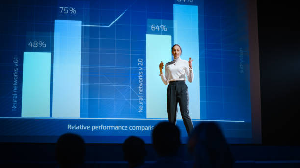 istock On Stage, Successful Female Speaker Presents Technological Product, Uses Remote Control for Presentation, Showing Infographics, Statistics Animation on Screen. Live Event / Device Release. 1226992793