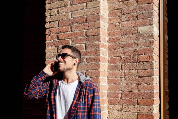 istock Young bearded man leaning on a bricked wall wearing sunglasses while using a smartphone outdoors. 1226993312