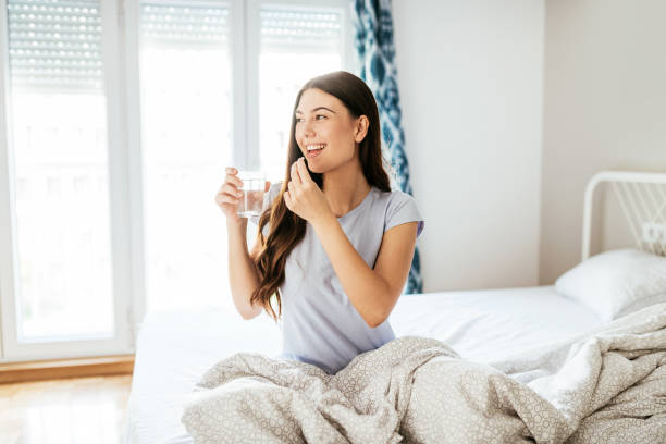 lächelnd eisern gesunde junge frau, die nahrungsergänzungsmittel und trinkwasser im bett - vitamin enriched stock-fotos und bilder