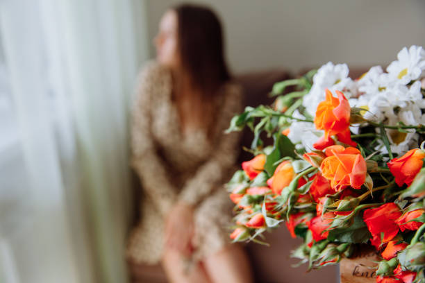 A bouquet of flowers: orange roses and white daisies lies in the front of the frame. In the background, a brunette girl is sitting and looking out the window.
