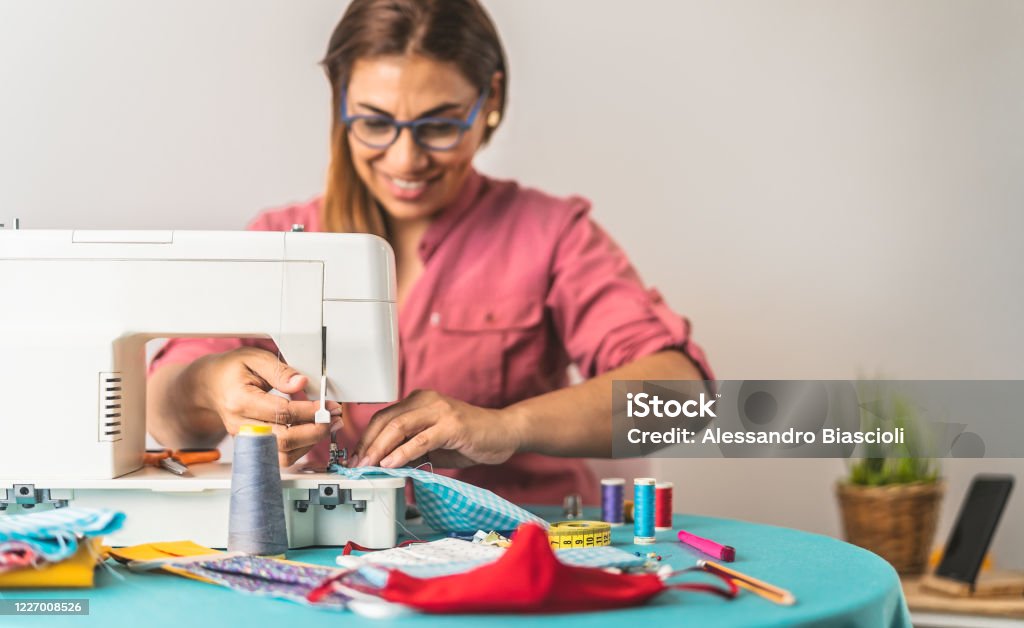 Happy Latin tailor seamstress woman sewing with machine homemade medical face mask for preventing and stop corona virus spreading - textile industry and covid19 healthcare concept Sewing Machine Stock Photo