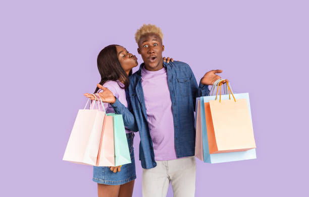 bella ragazza nera che bacia il suo fidanzato sorpreso con borse della spesa su sfondo lilla - couple black american culture kissing foto e immagini stock