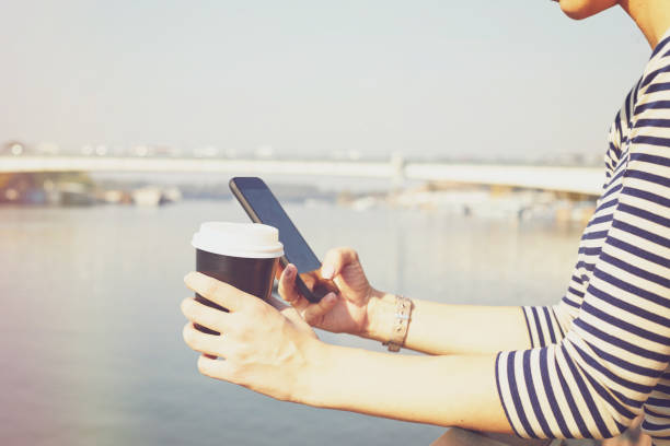 istock Beautiful woman using cellphone while drinking coffee near river. 1227012577