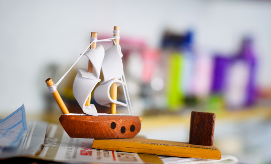 A wooden toy boat on a shelf with some papers under it, and other objects defocused on another shelf behind it.