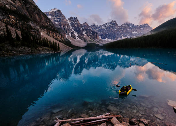 Young woman kayaks across mountain lake at sunrise Mountain range reflects in the blue water sunrise at moraine lake stock pictures, royalty-free photos & images