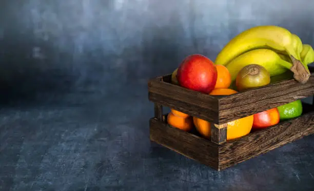 Photo of Wooden fruit box with banana, oranges, kiwi, peach, apricot on soft grey background with texture