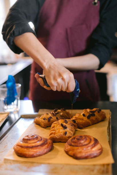 istock Vertical frame, baker's hands in process of squeeze maple syrup on freshly baked Danish buns lying on the counter of a local grocery store. Concept of daily routine at a fast food 1227021923