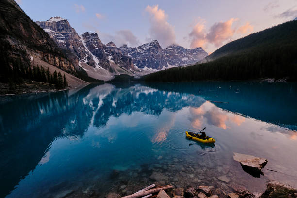 Young woman kayaks across mountain lake at sunrise Mountain range reflects in the blue water moraine lake stock pictures, royalty-free photos & images