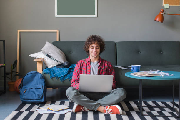Smiling College Student Sitting on the Floor and Using his Laptop Computer
