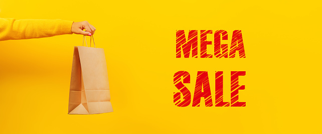 paper shopping bag in hand over yellow background, mega sale inscription, panoramic image