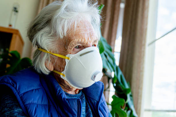 close-up shot of distraught elderly senior caucasian woman looking out the window feeling loneliness wearing an n95 protective face mask to prevent the spread of covid sars ncov 19 coronavirus swine flu h7n9 influenza illness during cold and flu season - flu virus cold and flu swine flu epidemic imagens e fotografias de stock