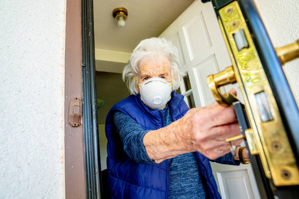 elderly senior caucasian woman cautiously opening the front door feeling unsure and wearing an n95 protective face mask to prevent the spread of covid sars ncov 19 coronavirus swine flu h7n9 influenza illness during cold and flu season - flu virus cold and flu swine flu epidemic imagens e fotografias de stock