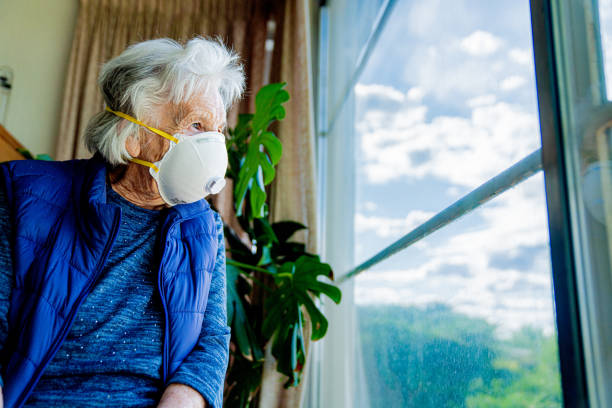 wide angle low shot distraught elderly senior caucasian woman looking out the window feeling loneliness wearing an n95 protective face mask to prevent the spread of covid sars ncov 19 coronavirus swine flu h7n9 influenza illness during cold and flu season - flu virus cold and flu swine flu epidemic imagens e fotografias de stock