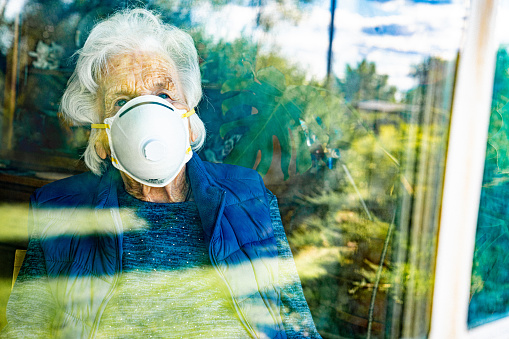 Distraught Elderly Senior Caucasian Woman Looking Out the Window Feeling Loneliness Wearing an n95 Protective Face Mask To Prevent the Spread of COVID SARS nCoV 19 Coronavirus Swine Flu H7N9 Influenza Illness During Cold and Flu Season