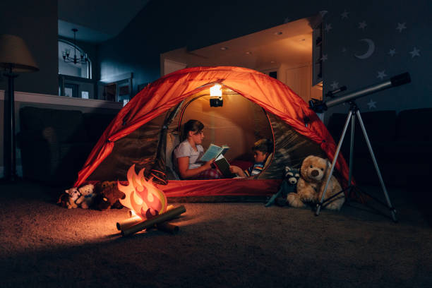 Girl and Boy Reading while Camping Indoors Two siblings, a brother and sister, camp inside their home due to the coronavirus restrictions and quarantine. They have pitched a tent along with their stuffed animal friends and have a fake campfire next to their telescope. They are reading books and making the best of their situation and long to return to the outdoors. staycation photos stock pictures, royalty-free photos & images