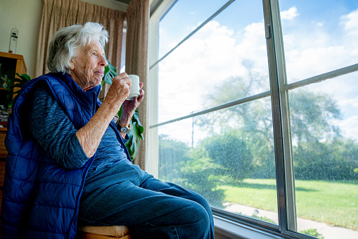 Elderly Senior Caucasian Woman Indoors Holding Coffee or Tea in a Mug and Looking Out the Window Enjoying the View in the Summer
