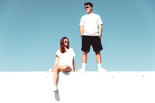 Young man and attractive young woman on concrete wall against the sky wearing sunglasses looking in opposite directions. Modern Urban Fashionable Street Style. Young Couple Concept Lifestyle Portrait.