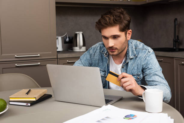 istock Selective focus of handsome man holding credit card and using laptop on kitchen table 1227040973