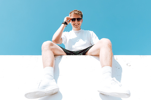 Happy smiling young man sitting on modern concrete wall, wearing sunglasses looking down with a happy smile. Shot from below against the blue sky. Urban Lifestly Youth Portrait.