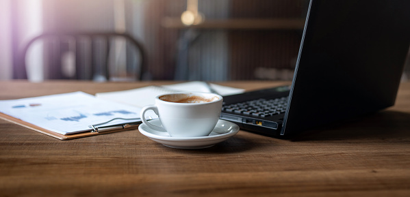 Close up white coffee cup on wooden desk with laptop and paperwork