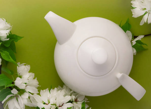 White tea pot, apple tree blossom branch, green background flat lay. Flowering buds top view template long web banner. Copy space backdrop mockup design. Floral spring frame flowers invitation concept