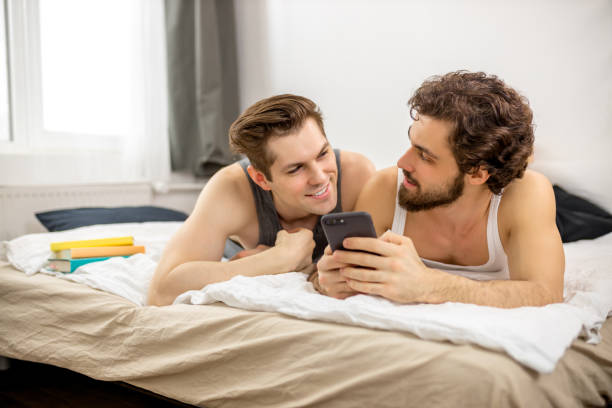 happy caucasian gay couple use smartphone positive same-sex couple relaxing in bedroom, looking at smartphone with excitement and smiling joyfully remembering sweet moments of last night party. homosexual men have leisure time together, lgbt community members unusual couple stock pictures, royalty-free photos & images