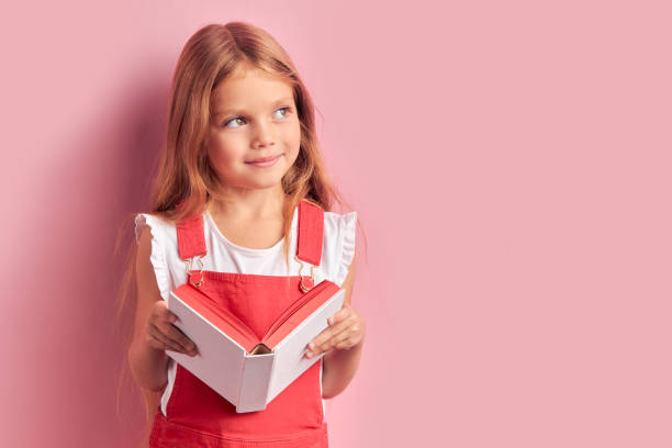 Portrait of dreamy coquettish girl with book in hands isolated over pink background Dreamy beautiful girl wearing red overalls looking side with book in hands, after reading interesting story cute girl stock pictures, royalty-free photos & images