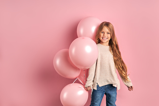 Smiling caucasian girl with long hair isolated over pink background, holding bunch of pink air balloons. The concept of children holiday birthday
