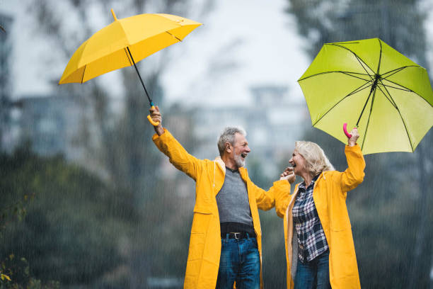 happy mature couple in yellow raincoats walking with umbrellas on a rainy day. - umbrella senior adult couple autumn imagens e fotografias de stock
