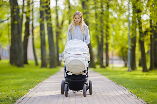 One young mother pushing white baby stroller and slowly walking at town green park in warm, sunny summer day. Spending time with infant and breathing fresh air. Enjoying stroll. Front view.