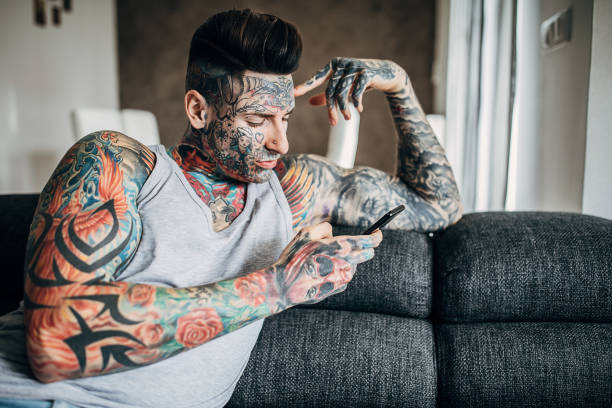 One young modern man with whole body covered in tattoos sitting on sofa in his living room and using smart phone.