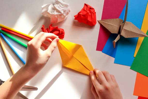 istock Children’s hands doing origami crane from yellow paper on white background with various school supplies. Step-by-step tutorial of origami. Concept of children's creativity, back to school. 1227093349