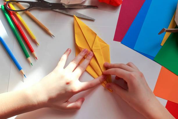 istock Children’s hands doing origami crane from yellow paper on white background with various school supplies. Step-by-step tutorial of origami. Concept of children's creativity, back to school. 1227093367