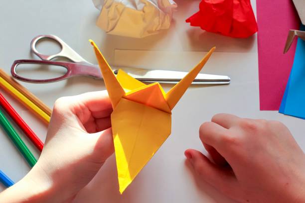 istock Children’s hands doing origami crane from yellow paper on white background with various school supplies. Step-by-step tutorial of origami. Concept of children's creativity, back to school. 1227093386
