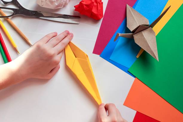 istock Children’s hands doing origami crane from yellow paper on white background with various school supplies. Step-by-step tutorial of origami. Concept of children's creativity, back to school. 1227093419