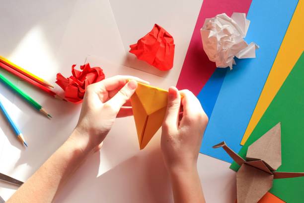 istock Children’s hands doing origami crane from yellow paper on white background with various school supplies. Step-by-step tutorial of origami. Concept of children's creativity, back to school. 1227093438