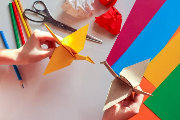 istock Children’s hands doing origami crane from yellow paper on white background with various school supplies. Step-by-step tutorial of origami. Concept of children's creativity, back to school. 1227093458