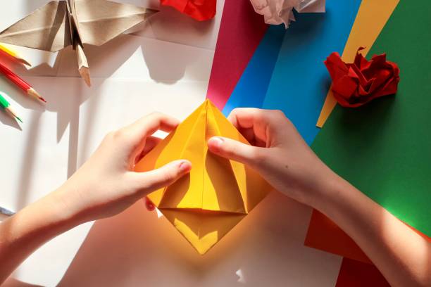 istock Children’s hands doing origami crane from yellow paper on white background with various school supplies. Step-by-step tutorial of origami. Concept of children's creativity, back to school. 1227093463