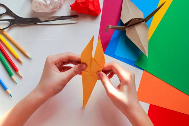 istock Children’s hands doing origami crane from yellow paper on white background with various school supplies. Step-by-step tutorial of origami. Concept of children's creativity, back to school. 1227093487