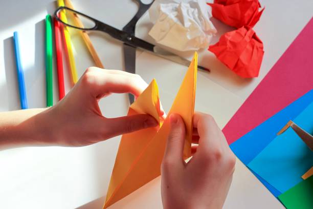 istock Children’s hands doing origami crane from yellow paper on white background with various school supplies. Step-by-step tutorial of origami. Concept of children's creativity, back to school. 1227093509