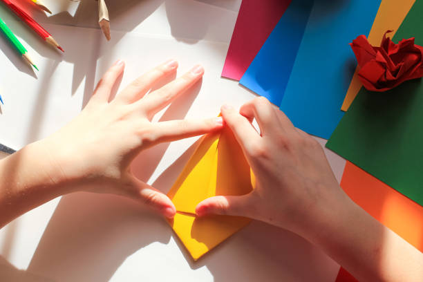 istock Children’s hands doing origami crane from yellow paper on white background with various school supplies. Step-by-step tutorial of origami. Concept of children's creativity, back to school. 1227093783
