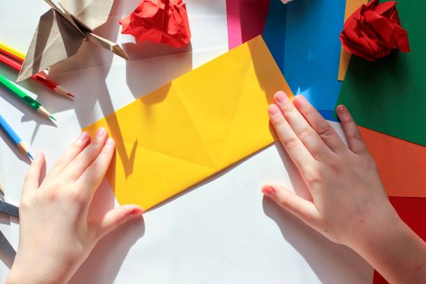 istock Children’s hands doing origami crane from yellow paper on white background with various school supplies. Step-by-step tutorial of origami. Concept of children's creativity, back to school. 1227093796