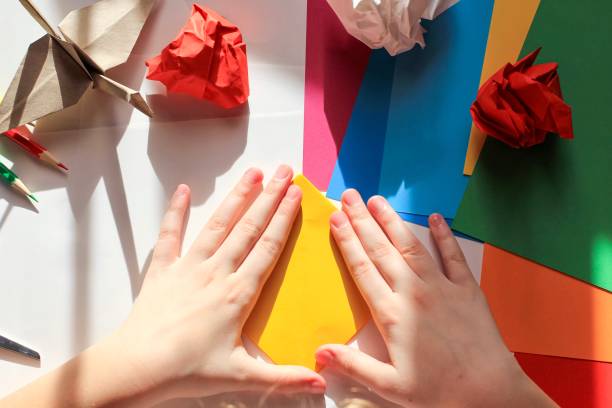 istock Children’s hands doing origami crane from yellow paper on white background with various school supplies. Step-by-step tutorial of origami. Concept of children's creativity, back to school. 1227093801